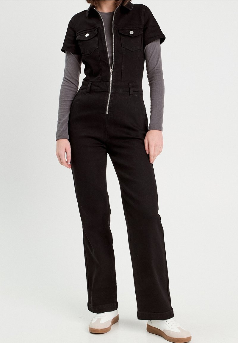 Černý džínový overal s krátkými rukávy, předním zipem a dvěma kapsami na hrudi. Kombinován se šedým tričkem s dlouhým rukávem a bílými teniskami.
