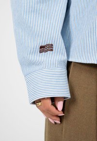 Blau-weiß gestreiftes Hemd mit einem Manschette, die ein braunes gesticktes Muster zeigt. Weicher Stoff, saubere Nähte und strukturierte Designdetails.