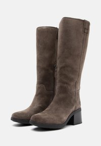 Cizme din suede maro, până la genunchi, cu toc chunky și fermoar lateral. Vârf rotund și textură netedă, având detalii de cusătură subtile.