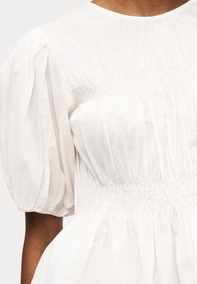 Blouse blanche avec manches courtes bouffantes, taille froncée et détails de tissu plissé sur la poitrine et les manches.