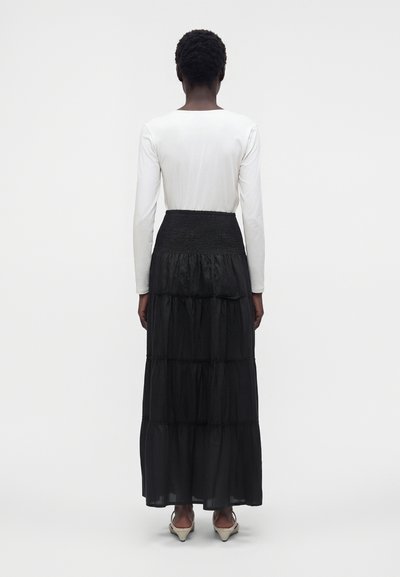Femme aux cheveux courts et bouclés, portant une longue jupe noire à volants, un haut blanc à manches longues et des chaussures à talons beige, debout, vue de dos.