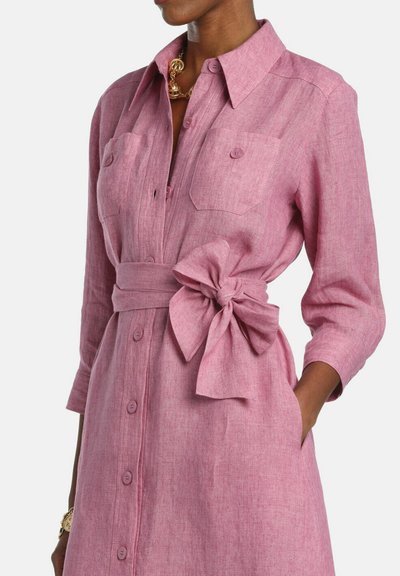 Frau trägt ein langärmeliges, rosa Knopf-Kleid mit Brusttaschen und einem großen, gebundenen Schleifengürtel an der Taille, steht mit einer Hand in der Tasche.