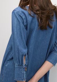 Denim-paita keskisinisessä, pitkällä hihalla, nappikoristeilla hihansuissa ja sivuleikkauksilla. Kankaan pinta on teksturoitu ja hieman haalistunut.