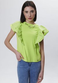 T-shirt in cotone verde neon con maniche arricciate e scollatura decorativa a catena. Indossato con jeans blu e presenta un'etichetta nel orlo.