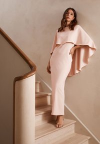 Długa różowa suknia z płynącą nakładką w formie peleryny, dopasowana sylwetka, miękki materiał i rąbek sięgający podłogi, zestawiona ze srebrnymi sandałami na paski.