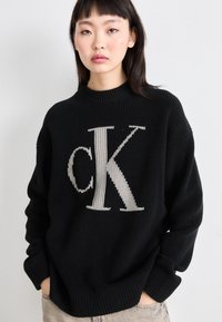 Črn pleteni pulover z velikim svetlo sivo "CK" logotipom spredaj, rebrastim ovratnikom in manšetami ter sproščenim krojem.