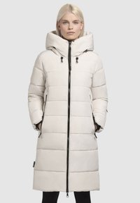 Manteau long matelassé en beige clair, avec un col montant, une capuche ajustable, un zip noir sur le devant et des poches latérales avec un léger éclat.