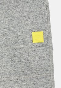 Pelēks treniņjakas materiāls ar faktūru. Iekļautas dzeltenas kvadrātveida etiķetes ar koka logotipu, kas atrodas kreisajā pusē.
