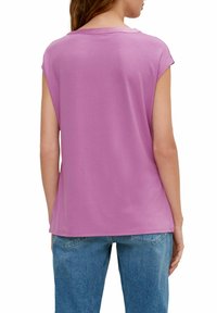 Kobieta w bezrękawnikowej lawendowej bluzce i niebieskich dżinsach, pokazana od tyłu na tle białego tła.