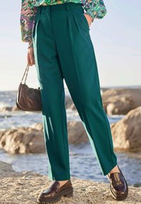 Groene high-waisted, geplooide broeken van soepel stof, gecombineerd met donkerbruin platform loafers en een kleine bruine handtas.