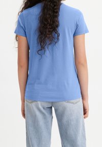 Kvinna med långtlockigt hår som bär en enkel blå kortärmad t-shirt och ljusblå denimjeans, vänd bort från kameran mot en vit bakgrund.