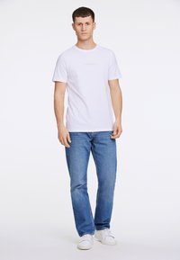 Valkoinen puuvillapaita, jossa on pyöreä kaula-aukko, sisältäen hienovaraisen harmaan logon. Yhdistettynä sinisiin farkkuihin ja valkoisiin lenkkareihin. Yksinkertainen ja rento muotoilu.