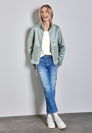 Veste bomber vert clair avec une texture pelucheuse sur les poches et le col, associée à un jean bleu usé et des baskets blanches.