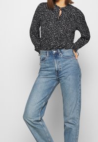 Blusa nera con motivo a macchie bianche, dettaglio a chiave sul collo, maniche lunghe; abbinata a jeans in denim azzurro chiaro a vita alta.