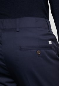 Námořnické kalhoty s hladkým povrchem, s kapsou na zadní straně s knoflíkem a malou značkovou visačkou, která ukazuje na propracované designové detaily.