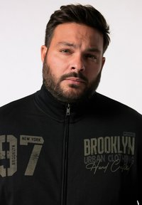 Black zip-up jacket with high collar; features printed text in gray: "BROOKLYN 97 NEW YORK" and "URBAN CLOTHING Hand Crafted."