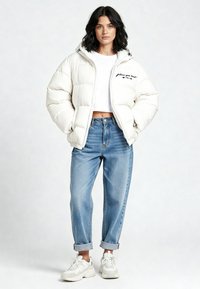 Veste blanche gonflée avec capuche, design court, et texte brodé noir. Associée à un jean bleu clair replié et des baskets blanches.
