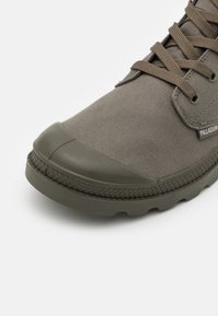 Zapatilla de lona gris con puntera de goma y suela texturizada, que presenta cordones redondos y una pequeña etiqueta de logo en el lateral.