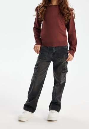 Enfant portant un haut à manches longues bordeaux, un pantalon cargo noir ample avec poches avant, et des baskets blanches à plateforme, debout devant un fond blanc.