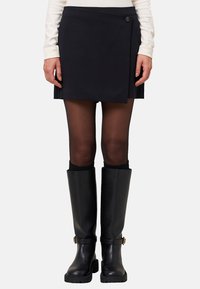 Femme portant une mini-jupe noire à manches, des collants noirs transparents et des bottes en cuir noir jusqu'aux genoux avec des boucles, se tenant contre un fond uni.
