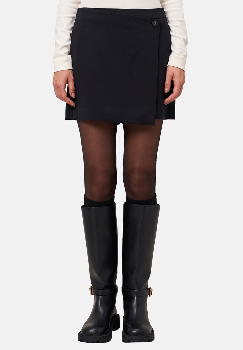 Femme portant une mini-jupe noire à manches, des collants noirs transparents et des bottes en cuir noir jusqu'aux genoux avec des boucles, se tenant contre un fond uni.