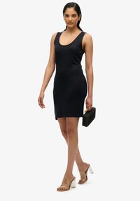 Robe sans manches bleu marine avec un décolleté arrondi, texture lisse et coupe ajustée. Le mannequin porte un sac à main rectangulaire noir et des sandales à talons transparents.