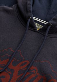 Navyblå hoodie med blød tekstur, der har snøre, et rødt grafisk design og en stribet inderside af hætten med et stofmærke.