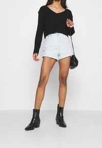 Schwarzer Strickpullover mit V-Ausschnitt, hellblaue hoch geschnittene Shorts und schwarze Knöchelstiefel. Das Outfit beinhaltet eine kleine schwarze Umhängetasche.