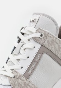 Gros plan sur une basket Michael Kors blanche et grise avec des empiècements texturés, des lacets blancs, un logo MK en métal sur la languette et le nom de la marque sur le côté.