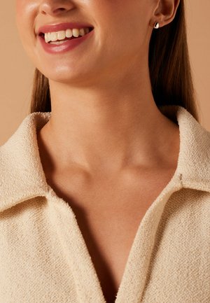 Lächelnde Frau trägt cremefarbene, strukturierte Jacke mit Kragen und kleine silberne und schwarze tropfenförmige Ohrringe vor beigem Hintergrund.