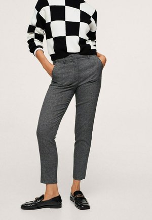 Person wearing black and white checkerboard sweater, gray slim-fit ankle pants, and black leather loafers with gold detail. Hands in pockets.