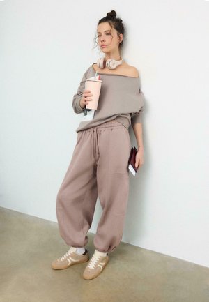Femme en pantalon de survêtement taupe ample, sweat-shirt décolleté, baskets beiges et casque autour du cou, tenant une tasse à boire et un portefeuille.