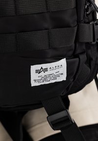 Μαύρο σακίδιο από νάιλον με αναγνωρίσιμο λογότυπο σε λευκό χρώμα, ρυθμιζόμενους ιμάντες και αξεσουάρ από ύφανση για επιπλέον επιλογές αποθήκευσης.