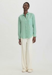 Helles grünes, langärmliges Hemd mit Knopfleiste und einer Brusttasche, kombiniert mit cremefarbenen weiten Hosen und hellen Sneakers. Weicher Stoff, lässiger Schnitt.