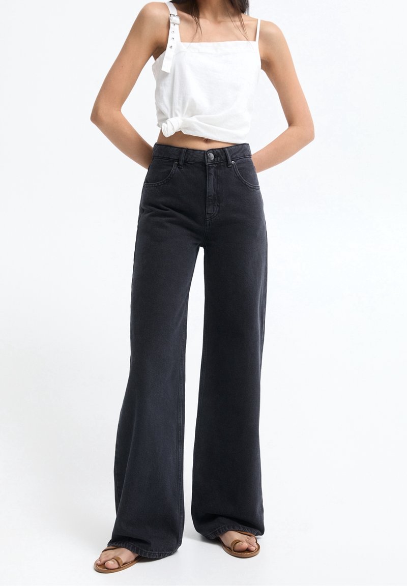 Jean flare taille haute noirs avec poches avant, coutures contrastées et une texture légèrement délavée, associés à un débardeur blanc noué.