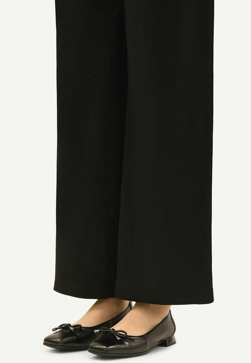 Pantalon large noir porté avec des ballerines en cuir noir avec de petits nœuds sur le dessus.