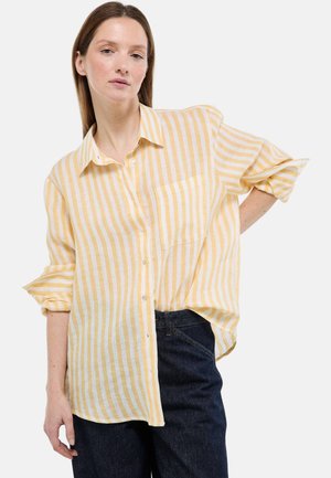 Femme portant une chemise à rayures verticales jaunes et blanches avec les manches retroussées et un jean bleu foncé, debout avec une main sur la hanche.