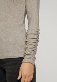 Långärmad beige topp med texturerade, rynkade ärmar, tillverkad av stickad tyg, i kombination med mörka jeans. Fokus på detalj av armen och ärmdesign.