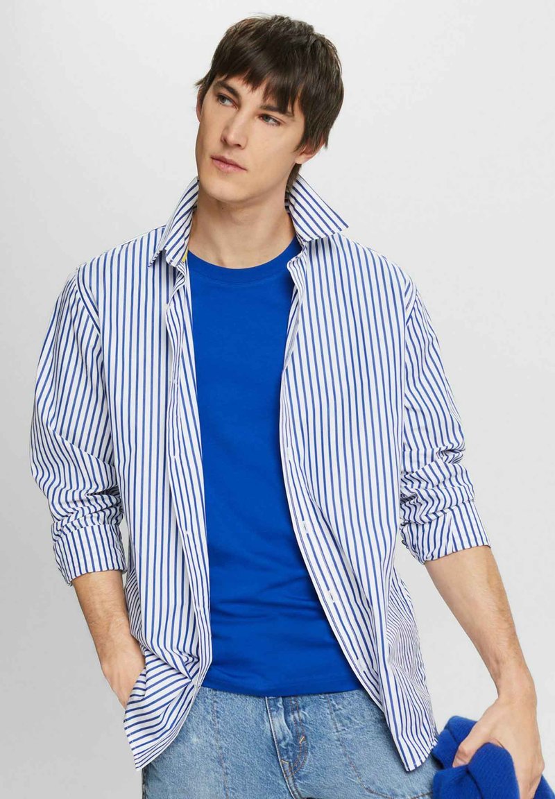 Skjorta med knappar i blå och vita vertikala ränder över en blå T-shirt med rund halsringning. Modellen bär ljusa denimjeans och håller i en blå föremål.