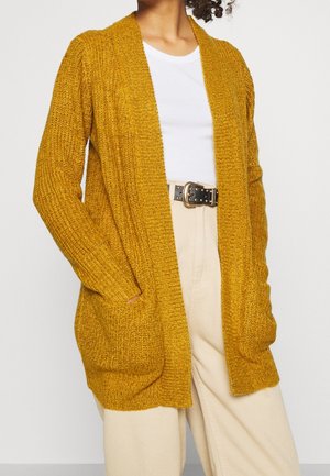 Femme portant un cardigan en maille jaune moutarde ouvert sur une chemise blanche, un pantalon beige taille haute et une ceinture noire avec une boucle dorée.