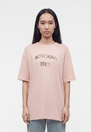 Femme aux longs cheveux noirs portant un t-shirt ample rose pâle avec le texte « MOSCHINO 1983 » et un jean bleu clair sur un fond blanc uni.