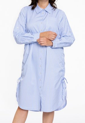 Vrouw draagt een losse blauw-witte verticale gestreepte overhemdjurk met lange mouwen en strikjes aan de zijkanten, met gekruiste armen.