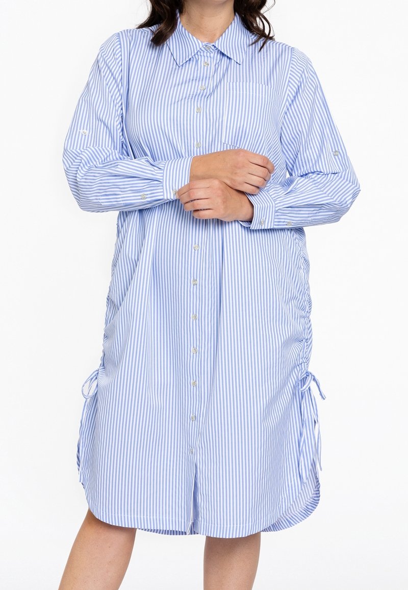 Femme portant une robe-chemise ample à rayures verticales bleues et blanches avec manches longues et liens sur les côtés, les bras croisés.