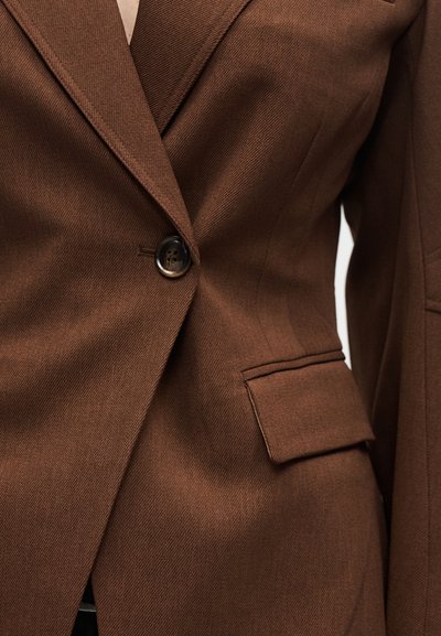 Κοντινό πλάνο καφέ ραμμένου σακακιού με γιακά με εγκοπές, ένα σκούρο κουμπί κλεισίματος και τσέπη με καπάκι στη δεξιά πλευρά.