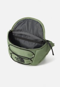 Sac de taille en tissu vert avec une ouverture zippée, intérieur gris et cordes élastiques externes pour un rangement supplémentaire. Équipé d'un patch logo à l'avant.
