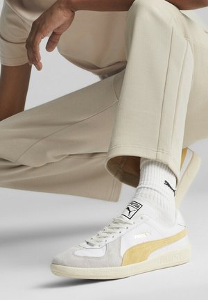 Puma sneakers met een bovenwerk van wit en beige suède, een gemarmerde zool en gouden accenten. Gecombineerd met lichtbeige broeken en witte sokken.