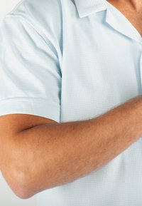 Chemise polo légère bleu clair à manches courtes avec un motif texturé, dotée d'un col décontracté et de poignets soigneusement finis, mettant en valeur un tissu doux.