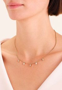 Ingrandimento di una persona che indossa una collana d'oro con cinque piccoli pendenti di cristallo rettangolari sulla pelle nuda.