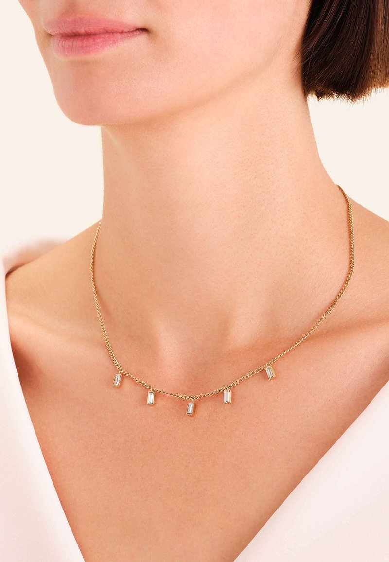 Ingrandimento di una persona che indossa una collana d'oro con cinque piccoli pendenti di cristallo rettangolari sulla pelle nuda.