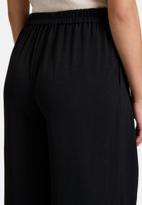 Zwarte wijde broek met een elastische tailleband en een gladde, textuurstof. De nadruk ligt op de achterkant en zijkant.